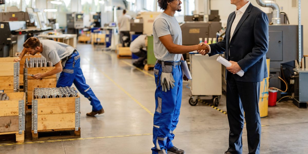 Happy black manual worker and engineer handshaking while greeting in industrial building.